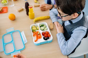 A young boy sits at a wooden table in school, enjoying a healthy lunch from a colorful lunchbox.