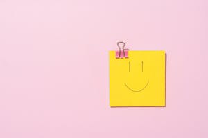 Yellow sticky note with a smiley face clipped to a pink surface, conveying happiness and simplicity.