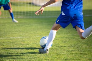A young soccer player in blue uniform kicks a ball towards the goal on a sunny day.